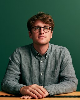 A young man in glasses seated at a desk, hands folded, against dark green.