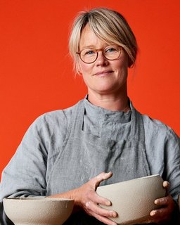 A potter in a linen apron holding two of her bowls, against flat red.