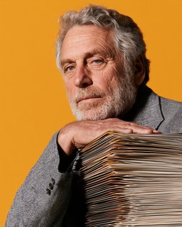 An older man leaning on a tall stack of papers, against amber.