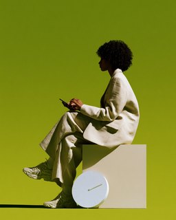A woman on a low block checking her phone, a wall clock propped beside her, against olive.
